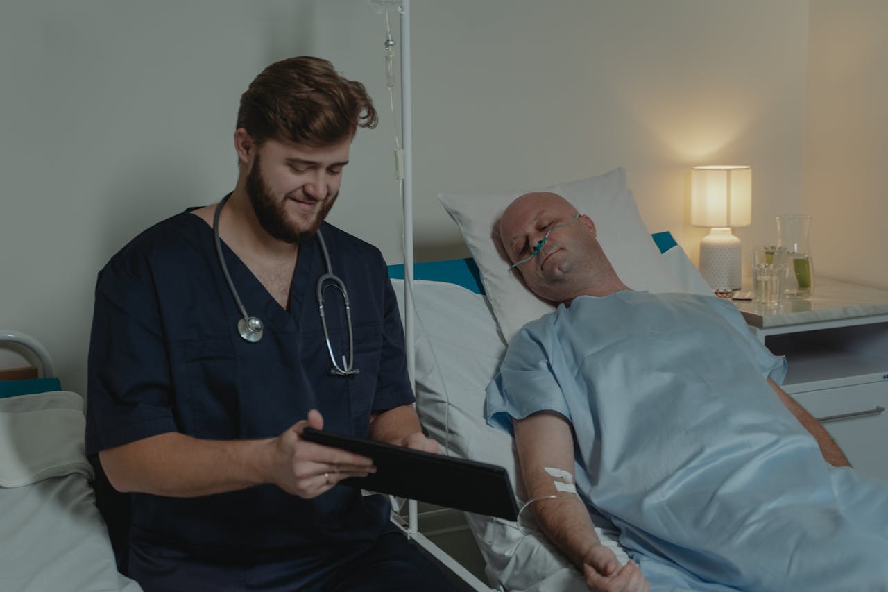 Doctor in scrubs using tablet to check health of patient lying in hospital bed with nasal cannula.